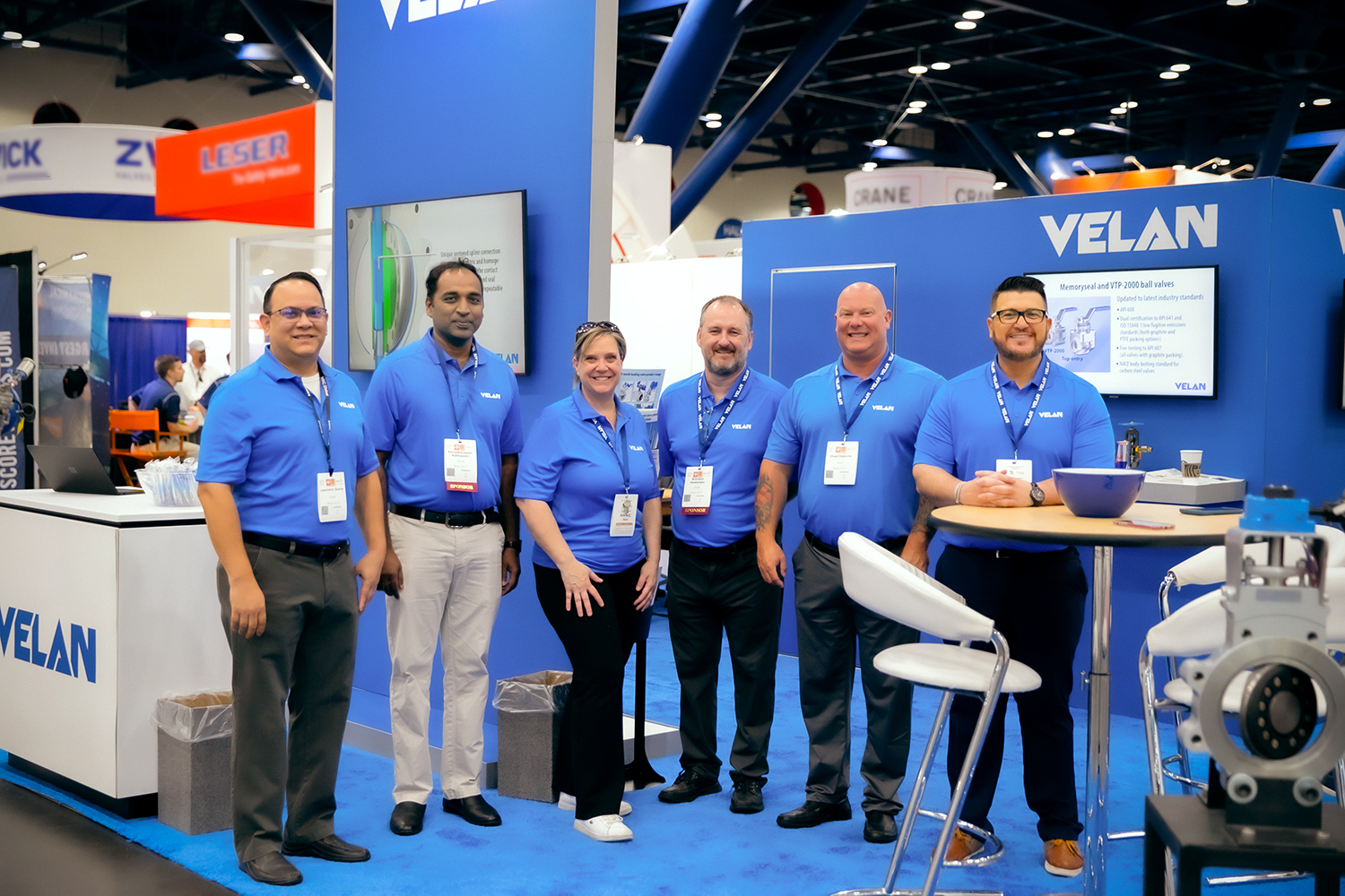A group of six people wearing matching blue shirts and name badges stand in a row, smiling at the camera. They are in an exhibition booth with the word "VELAN" prominently displayed on the walls. The booth has a table, chairs, and product displays in the background.
