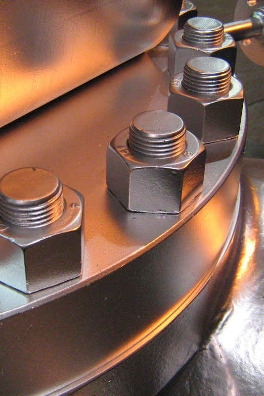 Close-up image of large metal bolts attached to a circular, industrial metallic surface. Warm light reflections highlight the contours of the bolts, creating a dramatic and detailed view of the mechanical components.