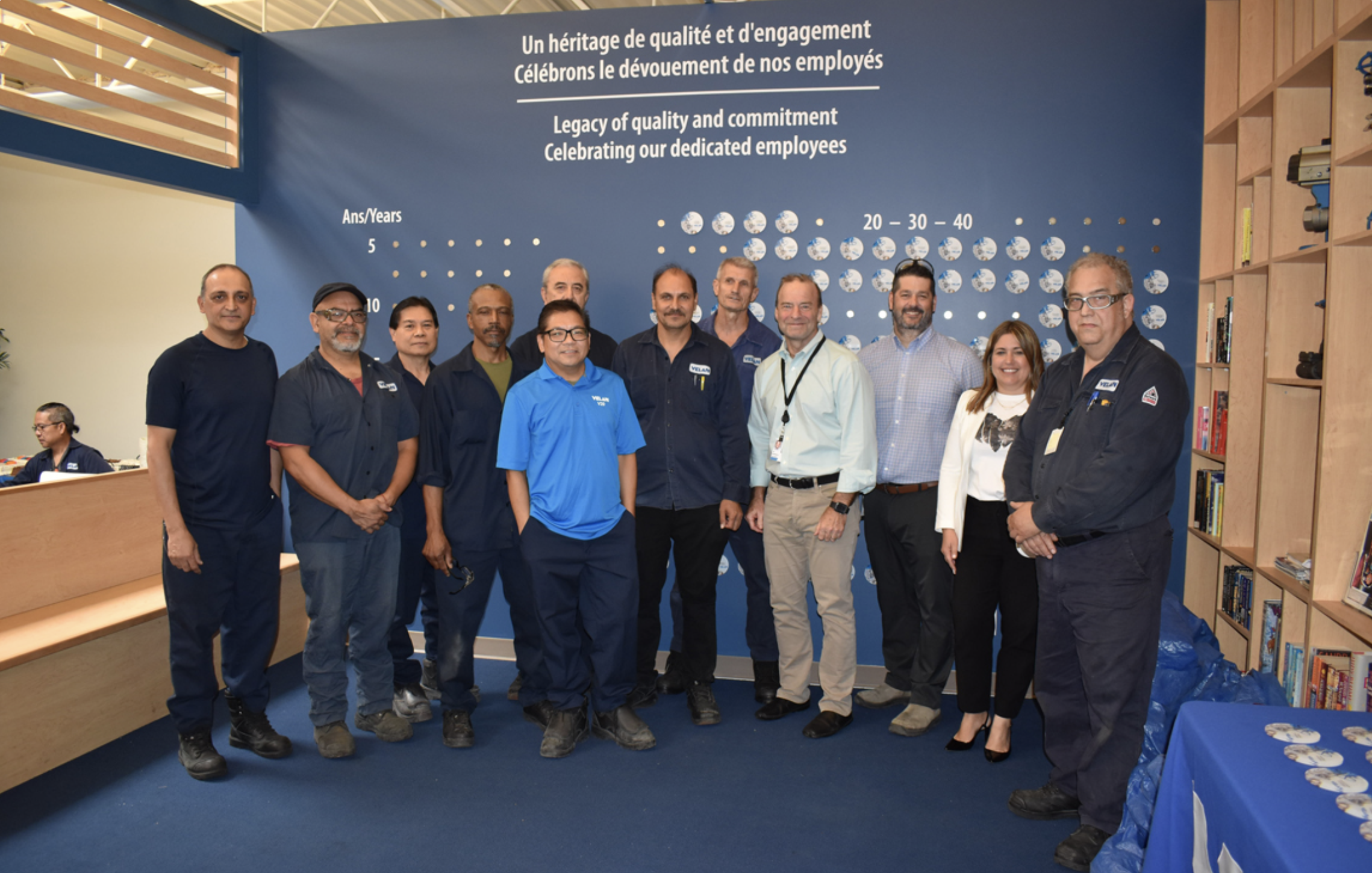 A group of eleven people stands in front of a blue wall adorned with white text celebrating employee commitment and quality. They are dressed in various professional and casual attire, smiling at the camera. A bookshelf and office setting are visible in the background.