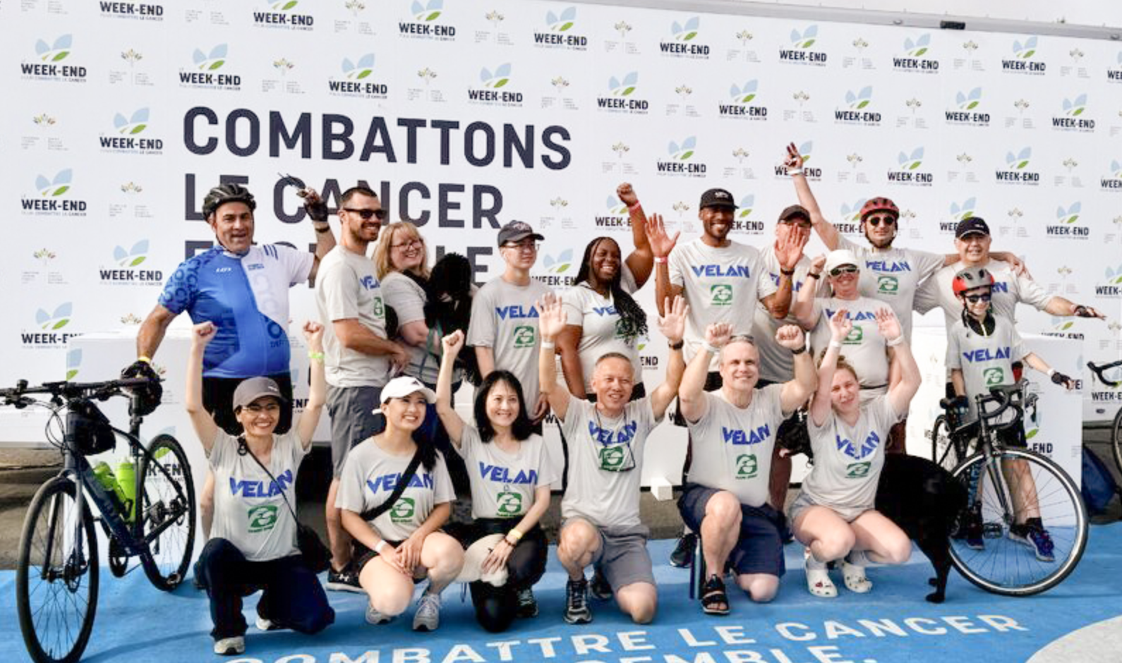 Group of individuals in matching shirts pose enthusiastically with bicycles in front of a backdrop reading "Combattre le cancer ensemble" (Fight cancer together). Some hold their fists up, and they seem to be celebrating or preparing for a cycling event.