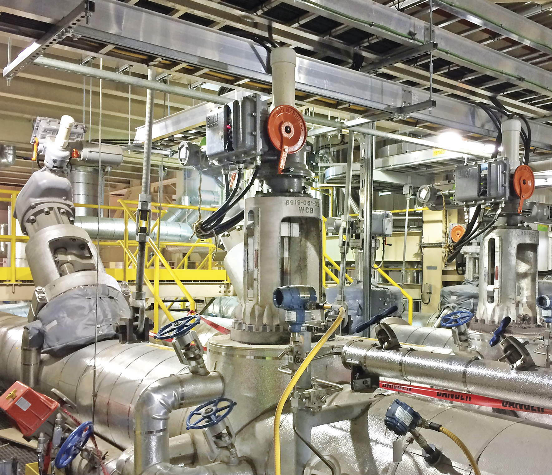 Industrial setting with complex machinery and pipes. Multiple valves and gauges are visible, including blue and orange wheel-handled valves. Overhead metal frameworks and conduits are part of the structure. Lighting fixtures hang from the ceiling, illuminating the area.