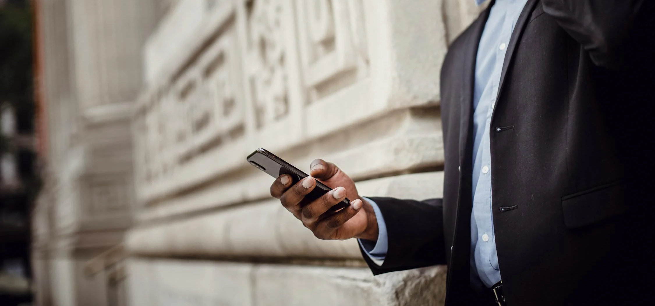 A person in a suit leans against a large, ornate stone building while talking on a smartphone. They are smiling and holding another phone in their other hand. The background is slightly blurred and shows part of the building's decorative stonework.